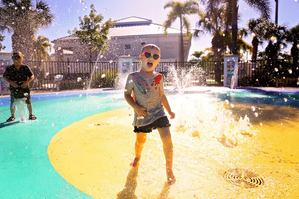 Child wearing heart-shaped sunglasses playing at splash pad in park.